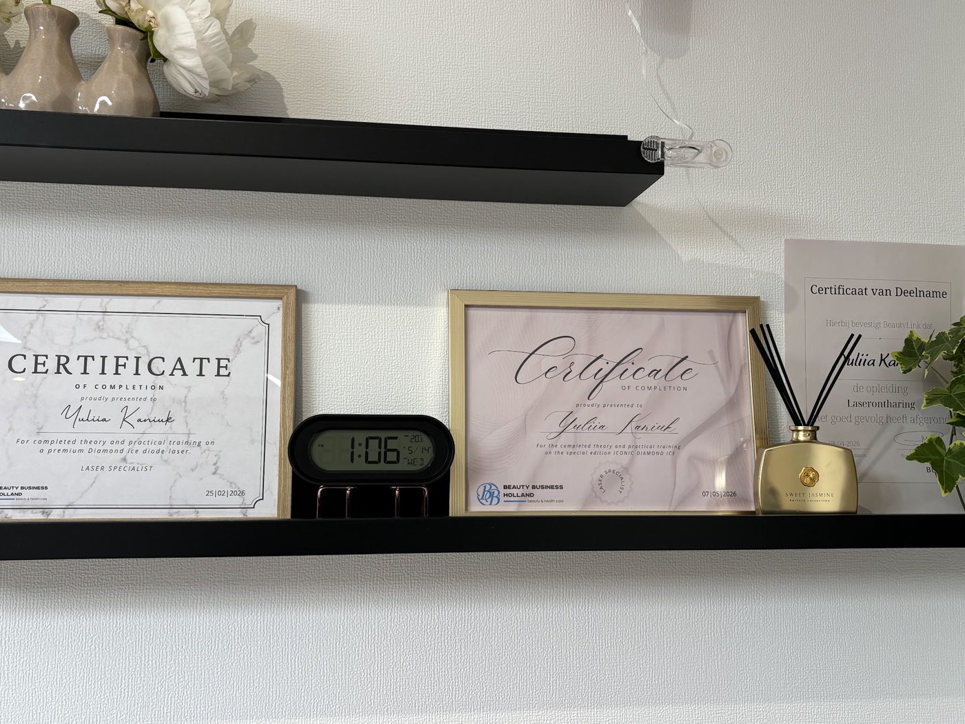 Twee ingelijste certificaten, een klokje en een geurstokjesflesje op een zwarte wandplank in de studio